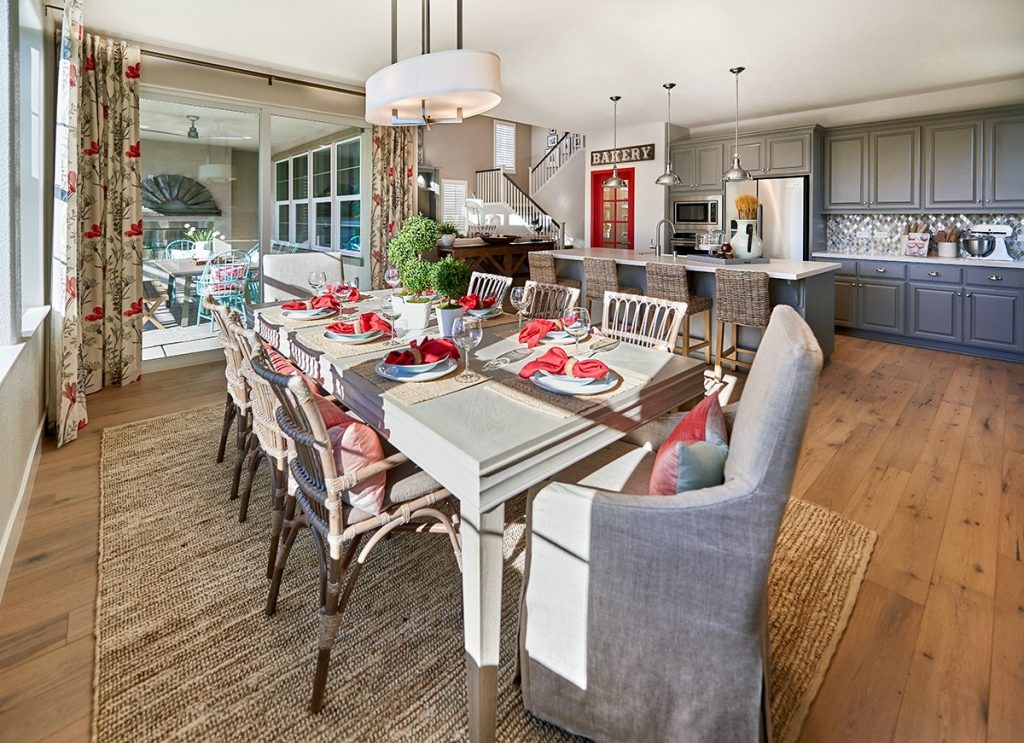 Dining room with off-white traditional dining table, wicker chairs and jute natural area rug, oval fabric and chrome chandelier, floral curtains, slider door, light brown wood flooring